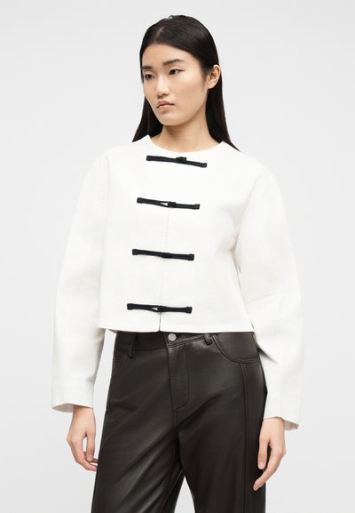 Γυναίκα με λευκή μπλούζα μακρυμάνικη με τέσσερα μαύρα κλεισίματα τύπου toggle και μαύρο δερμάτινο παντελόνι ψηλόμεσο, στέκεται μπροστά σε απλό φόντο.