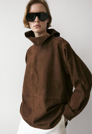 Persoon draagt een oversized bruine suède jas met hoge kraag en zwarte zonnebril tegen een effen lichte achtergrond.