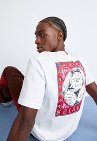 Hvid kortærmet t-shirt med en farverig fodboldgrafik på bagsiden, med stærke røde og blå accenter samt et detaljeret kantdesign.