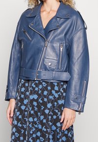 Kort blå skinnmotorcykeljacka med silverfärgade dragkedjor och detaljer, breda slag och midjebälte. Bärs över en blommönstrad klänning.