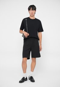 T-shirt e pantaloni corti in cotone nero abbinati a mocassini neri e calzini bianchi; il modello tiene una piccola borsa nera con tracolla regolabile.