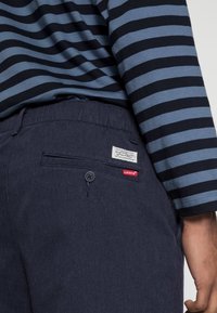 Levi's mörkblå byxor med en bakficka som har knappstängning och ett varumärkesmärke. Randig blå och svart långärmad skjorta synlig ovanför.