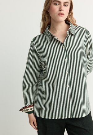 Woman wearing a green and white vertical striped button-up shirt with patterned cuffs, standing against a plain light background.