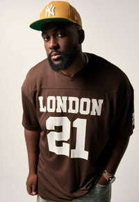 Homme portant un maillot de sport marron "London 21" et une casquette jaune moutarde des New York Yankees, regardant vers la caméra avec une expression neutre.