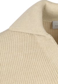 Beige gebreide trui, met een geribbelde textuur, een brede kraag en een klein label aan de binnenkant. De stof lijkt zacht en dicht.