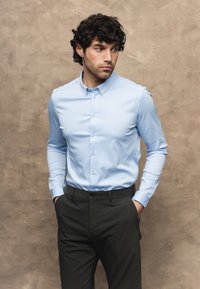 Uomo con capelli ricci e scuri che indossa una camicia azzurro chiaro abbottonata e pantaloni scuri, in piedi con le mani nelle tasche, guarda di lato.