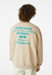 Felpa beige con colletto a girocollo, caratterizzata da testo stampato turchese sul retro che include "MELROSE AVENUE" e "Benvenuti in California".