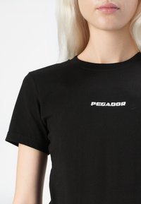 Fekete pamut póló, rövid ujjakkal és kerek nyakkivágással, fehér "PEGADOR" hímzéssel a mellrészen.