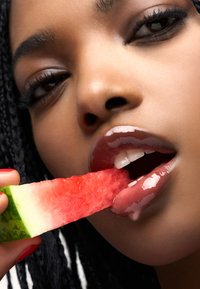 Zbliżenie osoby trzymającej kawałek arbuza, z połyskującymi ustami i warkoczykiem, na których widoczny jest czerwony lakier do paznokci.