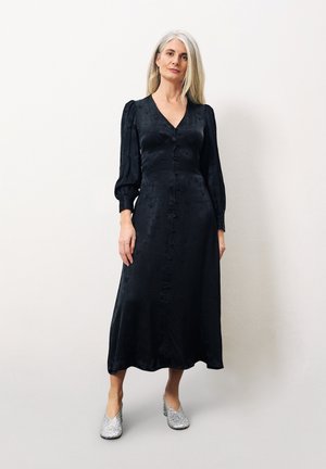 Zwarte midi-jurk met V-hals en pofmouwen, voorzien van knopen aan de voorkant. Gepaard met zilveren glinsterende hakken. Soepele textuur.