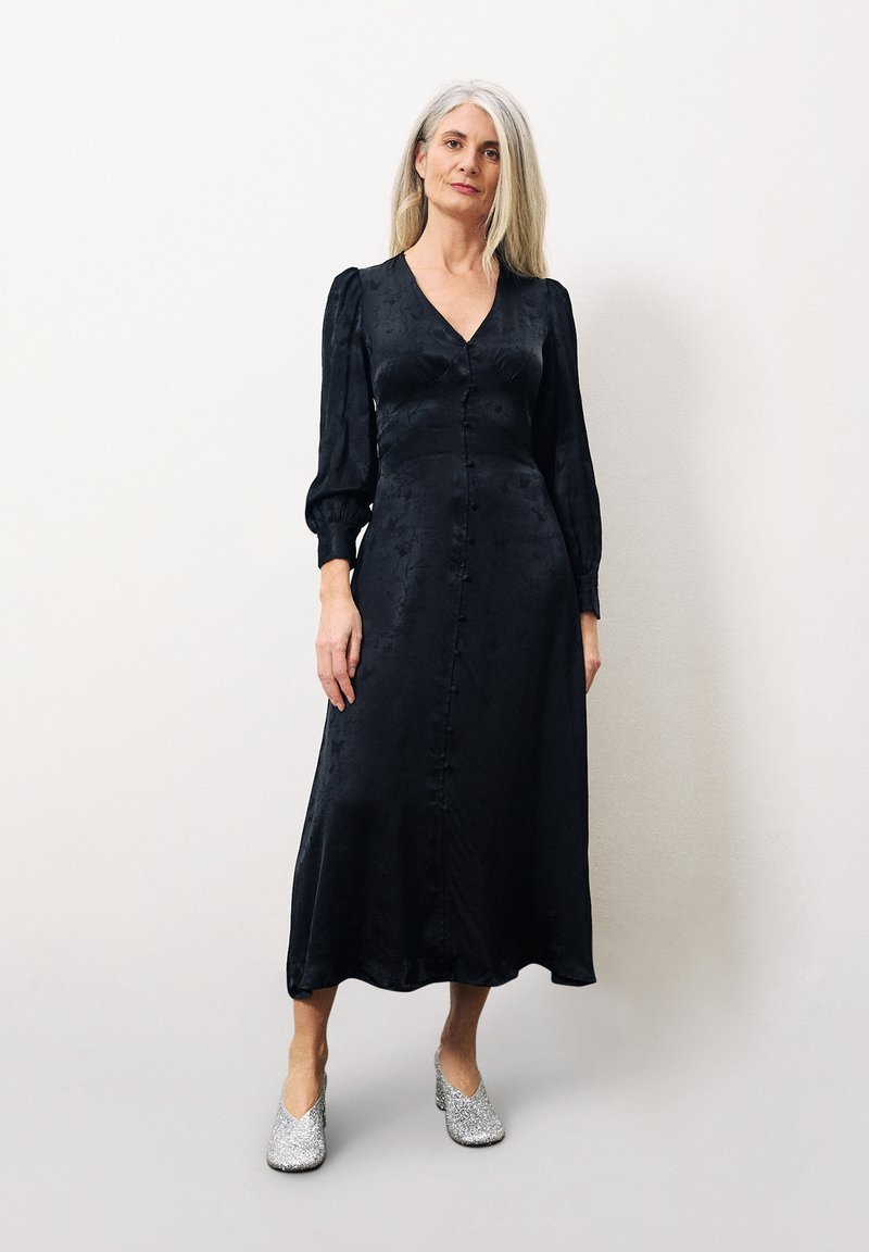 Schwarzes Midikleid mit V-Ausschnitt und Puffärmeln, mit Knöpfen an der Vorderseite. Kombiniert mit silbernen Glitzer-Absatzschuhen. Glatte Textur.