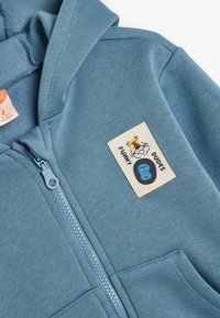Sudadera azul con cremallera hecha de una tela suave. Presenta un bolsillo tipo canguro y un parche con una cara de dibujos animados que dice "TIPOS GRACIOSOS" y una letra "B" azul.