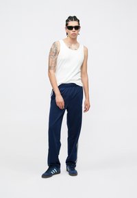 Camisola branca sem mangas, calças de treino azul-marinho com riscas brancas nas laterais e sapatos desportivos azuis. Pessoa usa óculos de sol escuros e tem cabelo trançado.