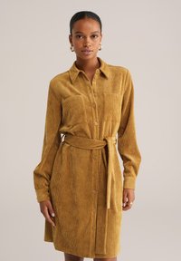 Rochie din catifea de culoare maro, cu guler, închidere cu nasturi și talie legată. Mâneci lungi cu mansete și două buzunare pe piept. Material texturat.