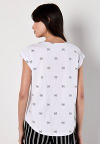 Una mujer con pelo largo de espaldas, vestida con una camisa blanca con pequeños patrones de lazo negro, combinada con pantalones a rayas.