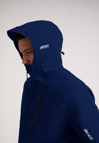 Marineblå vandtæt jakke med hætte, har lynlåslomme i front, justerbar snor og logo trykt på brystet og ærmet.