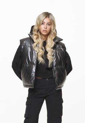 Schwarze Puffer-Weste mit glänzender Textur, übergroßem Kragen und Reißverschluss, getragen über einem schwarzen Sweatshirt und kombiniert mit schwarzen Cargo-Hosen.