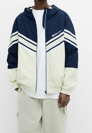 Person iført en marineblå og cremefarvet lynlåstrøje med hvide striber og teksten "UNION CITY", hvid skjorte og lyse beige cargobukser.