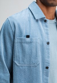 Man wearing light blue corduroy jacket with black buttons and chest pocket over a matching blue t-shirt.