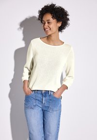 Top giallo chiaro a maniche lunghe con collo rotondo, vestibilità rilassata e tessuto testurizzato. Abbinato a jeans blu a vita alta con tasche frontali.