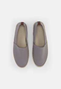 Espadrilles en tissu gris doux avec une semelle en jute tissée, présentant un accent en cuir marron au niveau du talon. Design minimaliste avec une construction à enfiler.