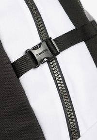 Tissu de sac à dos noir et blanc avec une fermeture éclair centrale, renforcé par des éléments en matériel noir et une boucle en plastique étiquetée "Superdry."
