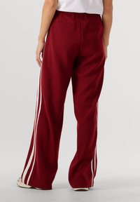 Een persoon in losse bordeauxrode broek met witte zijstrepen en witte sneakers, staand tegen een effen achtergrond.