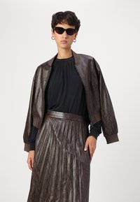 Braune Schlangenleder-Bomberjacke mit geripptem Kragen, kombiniert mit einer schwarzen Bluse und einem metallisch gefalteten Rock. Das Modell trägt schwarze Katzenaugen-Sonnenbrillen.