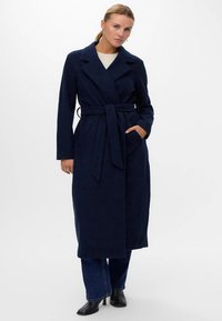 Cappotto lungo blu navy con vita sottolineata da una cintura, ampie rever e tasche laterali. Il tessuto appare texturizzato e caldo, abbinato a jeans scuri e stivaletti neri.