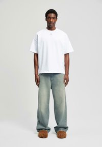 Uomo che indossa una t-shirt bianca oversize, jeans sbiaditi e larghi, pantofole marroni pelose e una collana d'argento, in piedi contro uno sfondo chiaro e semplice.
