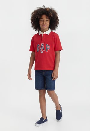 Garçon aux cheveux bouclés portant un polo rouge avec le texte "GAP", un short en jean et des chaussures sans lacets bleu marine, debout sur fond blanc.