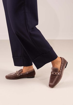 Mocasines de ante marrón con acentos de tachuelas, con punta redondeada y tacón bajo, combinados con pantalones anchos de color navy. Textura suave, diseño casual.