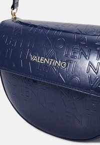 Lesklá námořnická modrá kabelka Valentino s embosovaným vzorem loga a název Valentino v zlatém kovovém provedení na předním klopě.