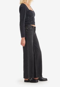 Czarna dżinsowa bluzka z długim rękawem i teksturowanymi bocznymi panelami, w połączeniu z czarnymi spodniami o wysokim stanie i luźnym kroju oraz płaskimi czarnymi butami.