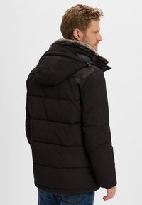 Man in een zwarte winterjas met vulling en een bontomrand kap, zijwaarts gekeerd, met blauwe jeans, op een witte achtergrond.