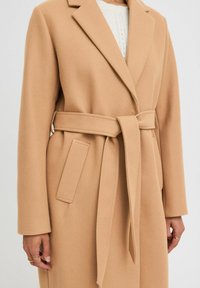 Manteau en mélange de laine couleur camel avec une taille ceinturée, des revers notés et des poches latérales. Tissu texturé avec une finition lisse.