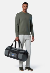 Mann trägt einen olivgrünen Pullover, hellgraue Hose und Sneaker und hält eine graue und schwarze Sporttasche mit einem runden Logo an seiner Seite.