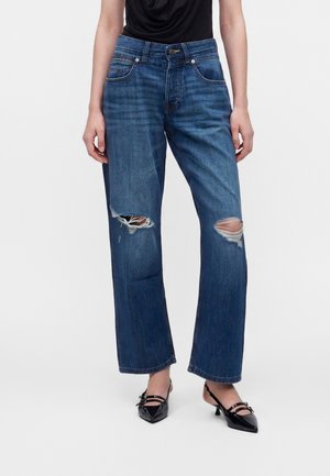 Person iført blå jeans med lige ben med revner ved knæene og sorte slingback-hæle med spids tå mod en ensfarvet baggrund.