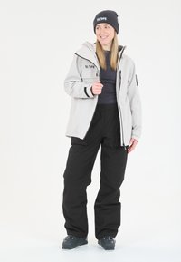 Tenue de ski comprenant une veste imperméable gris clair avec fermeture éclair et accents noirs, associée à un pantalon de neige noir et des bottes de ski grises.