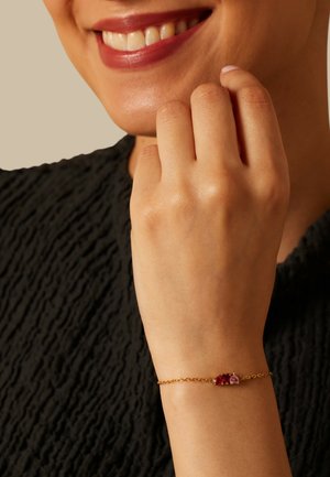 Glimlachend persoon met een zwart, textuur top, die een gouden armband met drie kleine rode en roze edelstenen om de pols draagt.