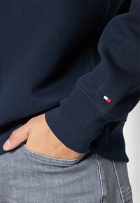 Granatowa koszula z długim rękawem z prążkowanym ściągaczem, z małym czerwono-biało-niebieskim logo na rękawie. Ręka spoczywa w szarej kieszeni z denimu.