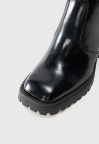 Bottine noire en cuir avec une finition brillante, semelle en caoutchouc noire épaisse, bout carré et détails de couture minimalistes.