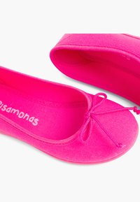 Ballerines en toile rose avec un bout arrondi, présentant un détail de nœud central et une surface en tissu lisse. Nom de la marque visible à l'intérieur.