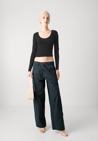 Svart ribbestrikket langermet crop topp kombinert med løse mørkegrønne bukser og en lys beige veske. Gjennomsiktige hælsandaler fullfører antrekket.