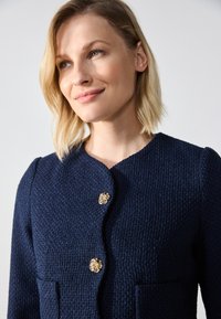 Veste texturée bleu marine avec un col rond, ornée de deux boutons floraux dorés et de deux poches avant. Finition en tissu lisse.