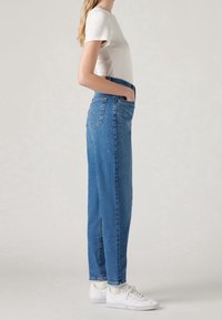Femme debout de profil portant des baskets blanches, un jean bleu taille haute avec une poche arrière, des chaussettes blanches et une chemise blanche à manches courtes.