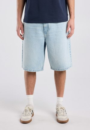 Man die lichtblauwe spijkershorts draagt, witte sneakers met zwarte details, witte sokken en een donkerblauw overhemd, staand op een effen achtergrond.