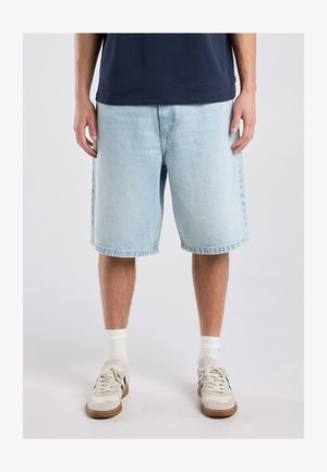 Man die lichtblauwe spijkershorts draagt, witte sneakers met zwarte details, witte sokken en een donkerblauw overhemd, staand op een effen achtergrond.