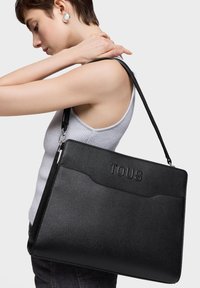 Bolso tote de cuero negro con logo "TOUS" en relieve, forma rectangular elegante, superficie texturizada y correa ajustable. Modelo lleva el bolso al hombro.