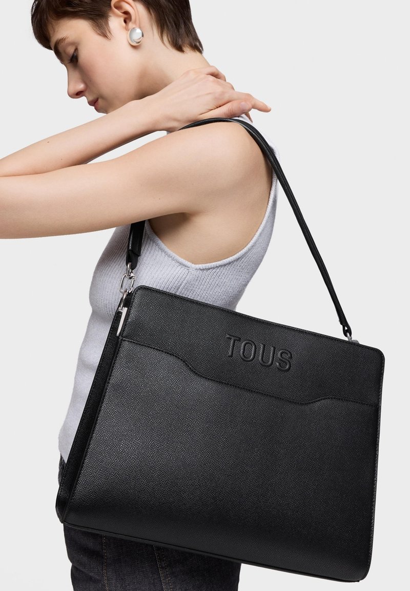 Bolso tote de cuero negro con logo "TOUS" en relieve, forma rectangular elegante, superficie texturizada y correa ajustable. Modelo lleva el bolso al hombro.